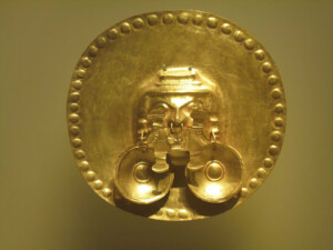 A large golden disc ornament decorated with intricate designs, featuring a face with prominent eyes and nose, and two circular, hanging elements attached at the bottom. Reflecting the rich history of mining in Latin America, the edge of the disc is adorned with raised dots.