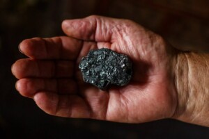 A hand is holding a piece of black coal, mined in Latin America. The coal has a rough, irregular surface with a slightly shiny texture. The background is blurred, focusing on the hand and the coal.