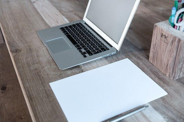 A silver laptop sits open on a wooden desk, next to a blank sheet of white paper. To the right of the laptop, there is a wooden pencil holder containing various writing utensils. The desk has a rustic, unfinished appearance, perfect for brainstorming ideas about doing business in Colombia.