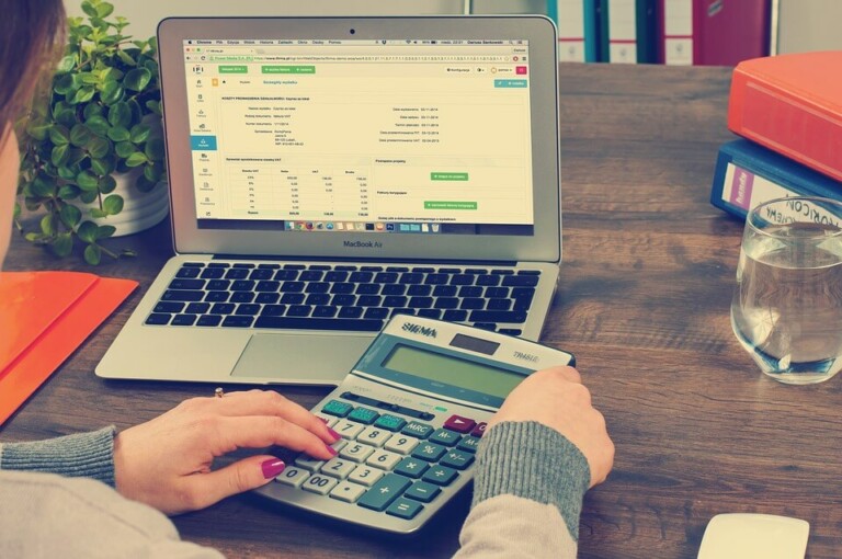 A person using a calculator at a wooden desk with a laptop open to a financial spreadsheet, possibly working on tax calculations. Also on the desk are a glass of water, a red and orange notebook, some documents, and a pen.