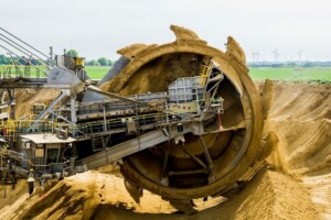 A large, industrial bucket wheel excavator is shown in operation at a mining site in Latin America. The machine's rotating wheel, fitted with multiple scoops, digs into the earth. In the background, wind turbines are visible on a distant, grassy horizon.