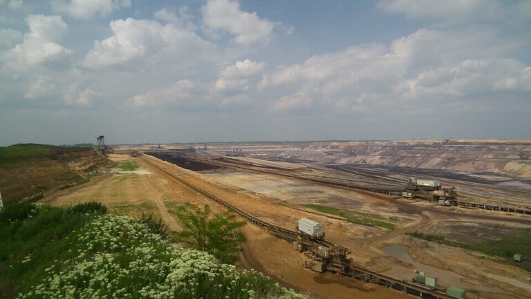 A large open-pit mining site under a cloudy sky. The scene features excavated dirt and layered earth, with mining equipment and conveyor belts typical of the mining sector visible. Green vegetation and small white flowers are in the foreground.