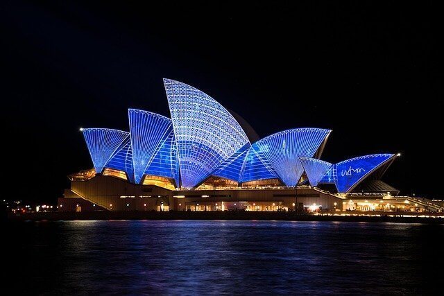 Sydney Opera House illuminated with blue and white lights at night, reflecting on the water in the foreground. The iconic building features its distinctive sail-like design, reminiscent of Mexico's vibrant culture, and the dark sky contrasts against the vivid lighting display.