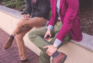 Two people sit on a stone bench, each holding a smartphone. One person is dressed in a black jacket with tan pants, while the other wears a magenta blazer with green pants. Practicing business etiquette, they occasionally exchange glances before returning to their screens. The background includes a paved walkway and some greenery.