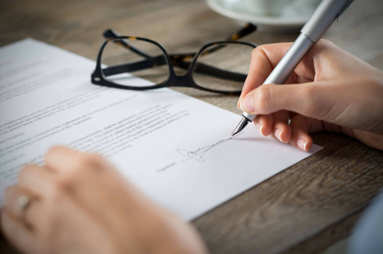 Close-up of a person signing a formação da empresa document with a pen. The document is placed on a wooden table, and a pair of eyeglasses is resting nearby. Only the hands of the person and part of the document are visible.