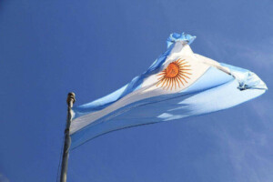 The image shows the flag of Argentina waving against a clear blue sky. The flag consists of three horizontal stripes of light blue, white, and light blue, with a sun emblem in the center of the white stripe, symbolizing optimism despite challenges to doing business in Argentina.