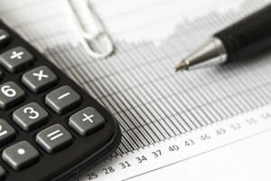 A close-up image of a calculator, a pen, and a paperclip on a piece of paper with a bar graph related to Chilean mining, accompanied by ruler markings. The calculator has visible buttons for addition and subtraction. The pen is positioned diagonally across the graph.