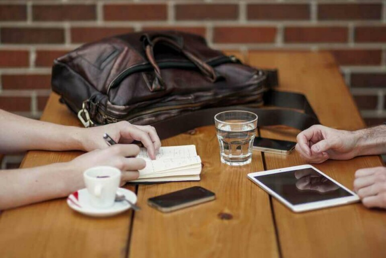 Two people sit at a wooden table with various items including a leather bag, a glass of water, a smartphone, and a tablet. One person writes in a notebook while the other’s hand rests near the tablet. A cup of espresso and Business English materials are also visible.