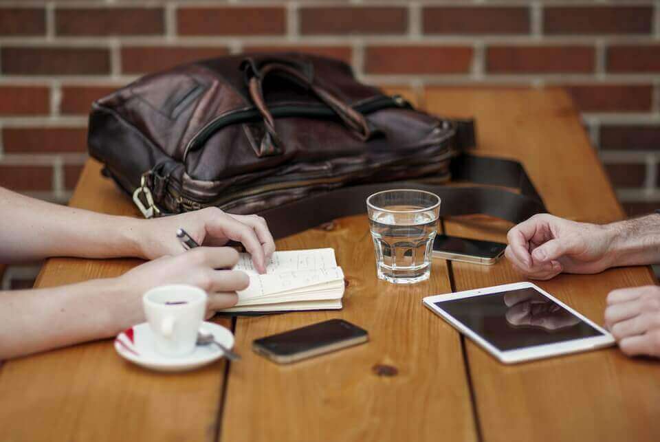 Duas pessoas sentadas em uma mesa de madeira com vários itens, incluindo uma bolsa de couro, um copo de água, um smartphone e um tablet. Uma pessoa escreve em um caderno enquanto a mão da outra descansa perto do tablet. Uma xícara de café expresso e materiais de inglês para negócios também estão visíveis.