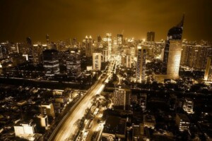 A city skyline at night with numerous lit buildings and a prominent building on the right with a spire-like top. The streets are illuminated by streetlights, and the scene has a sepia or amber-toned color filter giving it a warm hue, reflecting Jakarta's vibrant allure as we enter the Indonesian market.