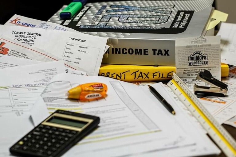 A cluttered desk covered with tax-related documents, a calculator, a spiral-bound book titled "Income Tax," highlighters, paper clips, and a receipt. There is also a notepad and various papers related to Formação da Empresa scattered around the work area.