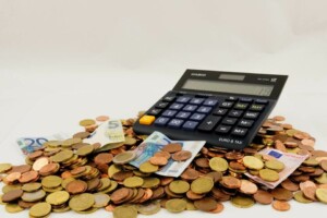 A calculator displaying numbers sits on a large pile of mixed currency, including euro coins and banknotes, symbolizing the complex process of VAT payments in Chile.