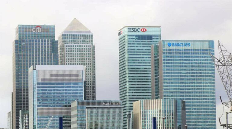 A cityscape showing a cluster of modern high-rise buildings, featuring prominent logos of Citi, HSBC, Barclays, and State Street. The buildings have mostly glass exteriors, reflecting the cloudy sky above—a true testament to the formation da empresa in the heart of the financial district.