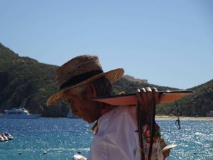 An elderly person wearing a straw hat and a white shirt holds a flat object with strings over their shoulder. Standing near water with a boat and mountainous landscape in the background on this sunny day, they embody the essence of sustainable development in Mexico.