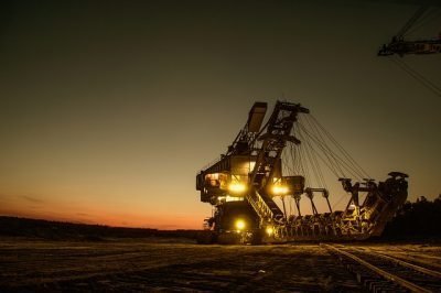 A large excavator with multiple mechanical arms operates in an open area at dusk, showcasing its prowess in the Colombian mining sector. The machine is illuminated by several lights, with the sky behind it showing hues of orange and blue as the sun sets.