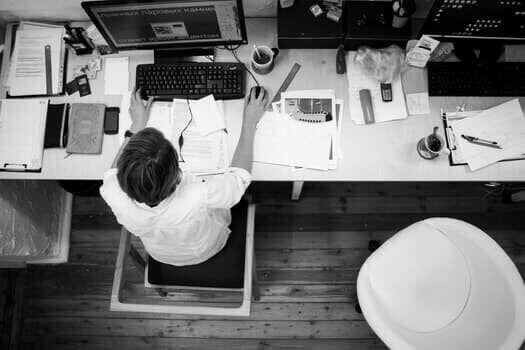 A black and white image of a person working at a desk with a computer and numerous papers, books, and stationery items spread out. The person is typing on the keyboard next to a cup of coffee. Among the scattered books is "Guía de Entrada al Mercado Latinoaméricano." The scene is viewed from above.