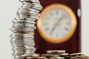 A precariously stacked pile of silver coins is in the foreground, with a classic wooden clock showing a blurred time in the background. The image underscores the balance between time and money, echoing themes of sustainable development in Mexico.