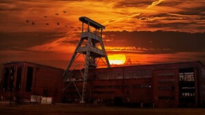 An industrial building stands against a dramatic sunset in Peru, with a tall metal structure in the center. The sky is ablaze with vibrant orange and red hues as birds fly in a V formation. This could be a future site for MINEM or APEC activities, but for now, the ground in front appears empty.