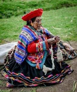 An elderly woman sits outdoors on the ground, dressed in traditional colorful garments and a red hat. She is holding and working with wool, possibly spinning it into yarn, as if weaving the intricate threads of her own company formation. Green grass is visible in the background.