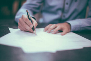 A person wearing a long-sleeved shirt is writing on a sheet of paper using a pen. The individual's right hand is holding the pen while the left hand rests on another document related to company formation. The background is blurry, drawing focus to the writing activity.