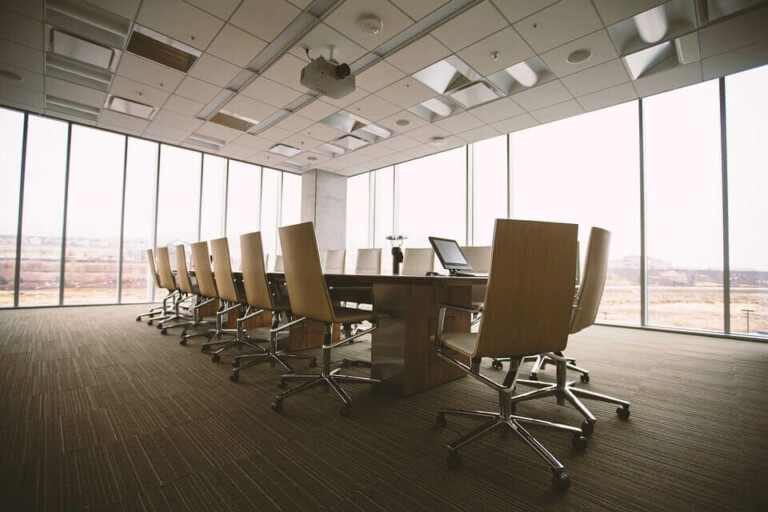 Una sala de conferencias grande y moderna con una larga mesa de madera rodeada por doce sillas de oficina con ruedas de color beige. Perfecta para cualquier aniversario corporativo, la sala cuenta con ventanales de piso a techo que permiten luz natural, iluminación cenital y un proyector montado en el techo.