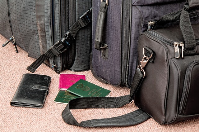 A close-up of travel bags and suitcases on a carpeted floor. Two passports, a wallet, and a small leather bag with a shoulder strap are visible in front of the larger suitcases. The items suggest preparation for a trip or exploring various visa types in Peru.