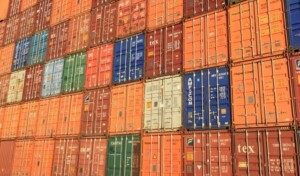 The image shows a large stack of multicolored shipping containers arranged in rows and columns within LATAM Free Trade Zones. The containers feature various logos, company names, and numbers, indicating different origins and destinations. The predominant colors are orange, blue, red, and yellow.