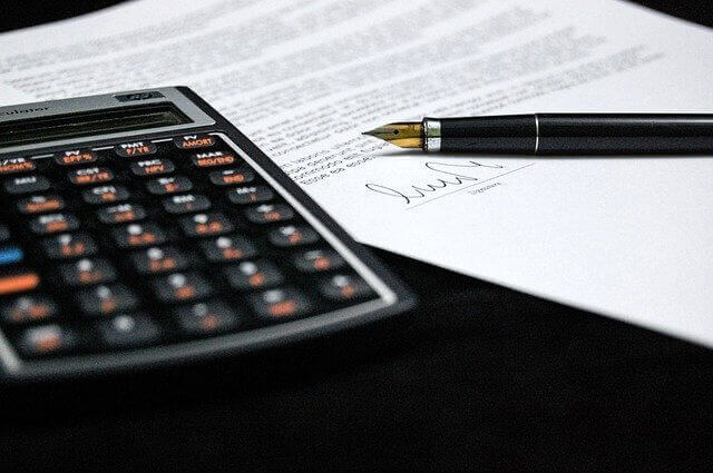 A calculator and a fountain pen rest on a document with printed text. The pen's cap is off, and the pen lies on the lower portion of the document, which pertains to Mexico International Trade and includes an illegible signature. The background is a dark surface.