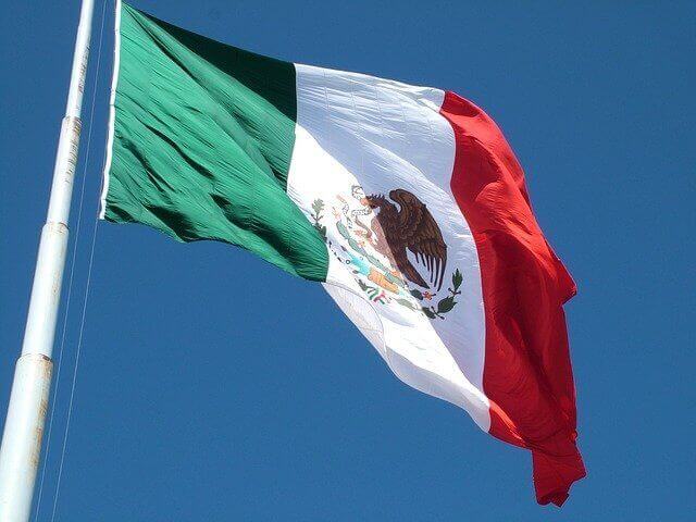 A flag featuring three vertical stripes of green, white, and red, with a coat of arms in the center of the white stripe, is hoisted on a pole against a clear blue sky. This iconic symbol of international business in Mexico includes an eagle perched on a cactus holding a snake.