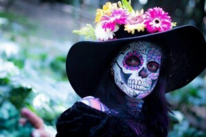 Person wearing intricate Day of the Dead face paint and a large black hat adorned with various colorful flowers. The background is slightly blurred, showing an outdoor setting with greenery, capturing the vibrant cultural essence that is often seen during international business events in Mexico.