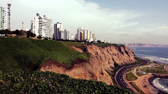 Un paisaje urbano costero presenta altos edificios blancos a lo largo de la cima de acantilados cubiertos de verde. Abajo, una carretera curva a lo largo de la costa. El mar es visible a la derecha, con suaves olas que llegan a la orilla bajo un cielo parcialmente nublado, ofreciendo un telón de fondo ideal para cualquier negocio en Perú.