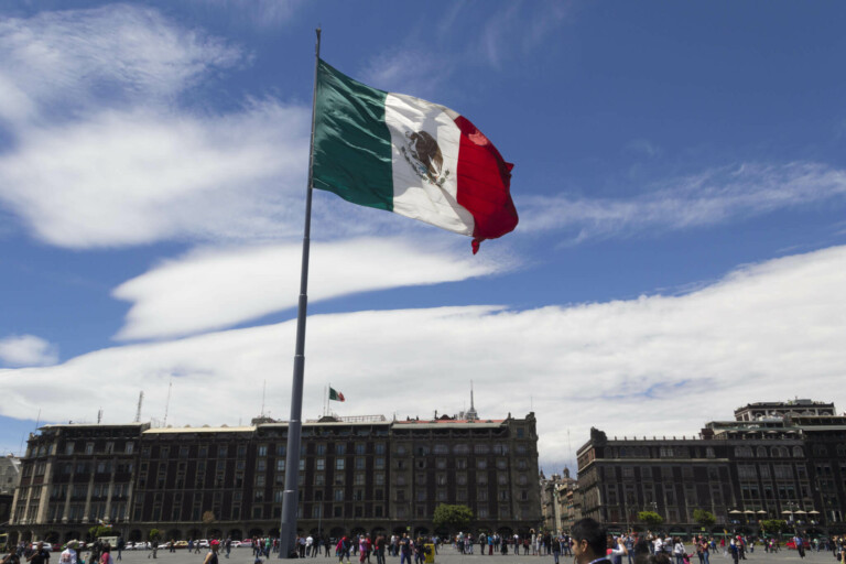 Una gran bandera mexicana ondea prominentemente en el aire en una plaza urbana bajo un cielo azul brillante con nubes dispersas. La gente se reúne alrededor de la plaza para celebrar un aniversario, mientras los edificios históricos se alinean al fondo.