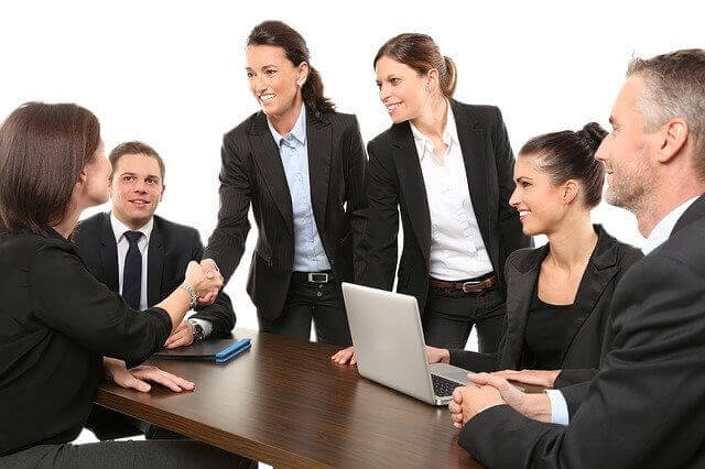 Five people in business attire sit around and stand near a table. Two of them are shaking hands while the others smile, discussing how to expand into Latin America. A laptop is open on the table, and the background is plain white.