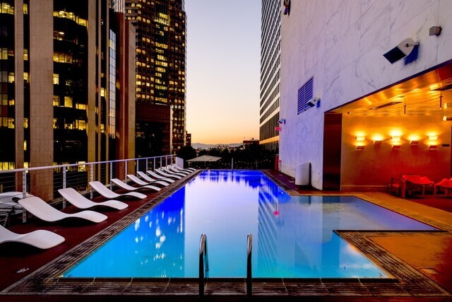 Zona de piscina en la azotea al atardecer, rodeada de edificios altos. La piscina tiene un pequeño tramo de escaleras que conducen al agua. Los sillones blancos están alineados a un lado y el área de la terraza está iluminada con una cálida iluminación naranja en el otro lado, capturando una de las últimas tendencias del mercado en comodidades de lujo.