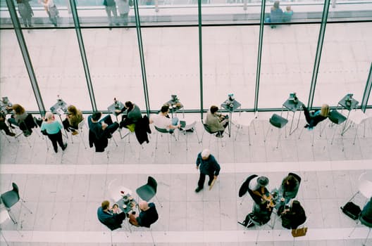 Una vista aérea de una moderna zona de estar interior con gente sentada en pequeñas mesas redondas. El espacio, indicativo de las tendencias del mercado, cuenta con altas paredes de vidrio. Las personas participan en diversas actividades, como comer, beber y conversar, mientras se ve a algunos individuos caminando.