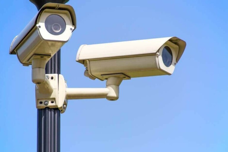 Dos cámaras de vigilancia blancas están montadas en un poste negro contra un cielo azul claro. Una cámara apunta hacia adelante y la otra se inclina ligeramente hacia abajo. Las cámaras tienen forma rectangular con carcasas protectoras, lo que permite vigilar atentamente el tráfico en Negocios Latinoamérica.