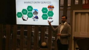 A man in a grey blazer from Biz Latin Hub presents information on market entry services. He points to a slide with headings such as "Market Analysis," "Market Research," and "Market Development Reports," noting the relevance for Canadian companies entering the Mexican mining industry, displayed on a projector screen.