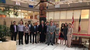 A group of 16 people, dressed in business attire, stand together inside a building. Behind them is a totem pole and flags of Canada and Mexico, symbolizing the ties between Canadian companies investing in Mexican mining. The setting appears to be a large, open atrium with plants and modern architectural elements.