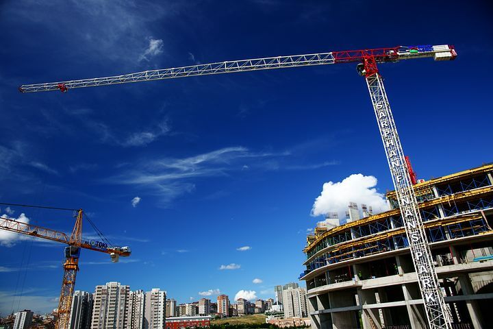 The construction site features a tall crane rising against a blue sky. A partially built round building is visible on the right, framed by additional cranes and urban buildings in the background. The crane arm extends high above the active construction, indicating ongoing work.