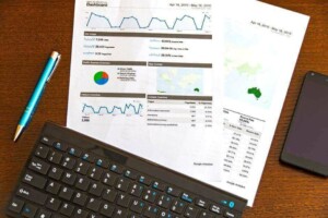 A desk with a piece of paper showing charts and graphs, a blue pen, a black keyboard, and a black smartphone. The paper includes data on website traffic and SAP S/4HANA trends, with segments and insights displayed over time.