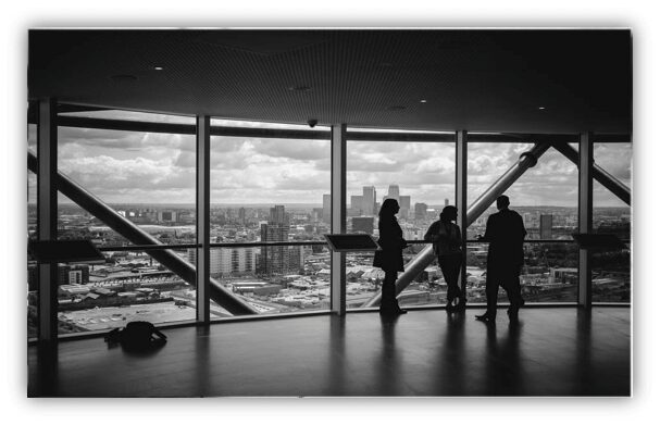 A black-and-white image showcasing three people standing near large windows at an observation deck. The cityscape, filled with buildings and a partly cloudy sky, is visible through the windows. The individuals appear to be engaged in conversation about accounting services.