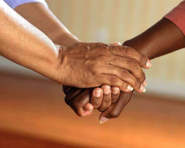 Two pairs of hands are shown in the image. One pair, with lighter skin, is clasping the other pair, with darker skin, in a supportive manner against a blurred background. It evokes thoughts of unity and strength amidst complex German relations and broader global issues.