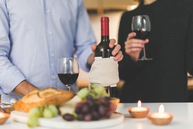 Two people are standing at a table, savoring glasses of red wine. The table, adorned with a wine bottle, lit candles, a loaf of bread, grapes, and nuts, reflects the richness of exporting Peruvian wine. One person pours more wine into a glass while the other holds theirs, ready to taste.