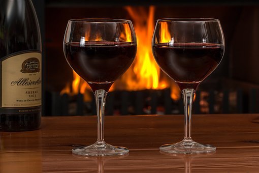 Two glasses of red Chilean wine are placed on a wooden table in front of a lit fireplace. A wine bottle is partially visible on the left side of the image. The bright flames in the background create a warm and cozy ambiance, showcasing an excellent trading opportunity for fine wines.