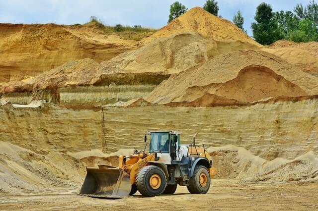 A large yellow and white bulldozer with a front shovel is positioned in a sandy quarry. Surrounding the vehicle are steep, layered sand and rock formations. Trees are visible in the background under a partly cloudy sky, highlighting Minería en Colombia's diligence in such landscapes.