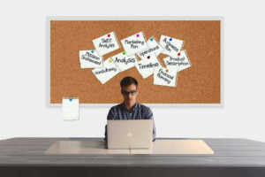 A person is sitting at a desk working on a laptop. In the background, there is a corkboard with numerous pinned notes labeled with business-related terms such as "SWOT Analysis," "Marketing Plan," and "Product Description." A blank notepad hangs to the left, hinting at potential strategies for business risk management in Argentina.