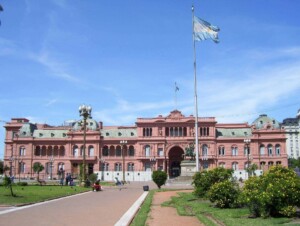 An image showing the Casa Rosada, the executive mansion and office of the President of Argentina, in Buenos Aires. The building is pink with ornate architectural details. In front, there is a large Argentine flag on a flagpole and a well-maintained garden, where discussions about leyes laborales often take place.