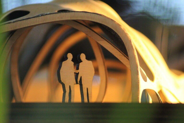 Two silhouette figures made of cardboard stand together under a wooden arch structure with circular cutouts, symbolizing the early stages of Formação da Empresa. The background is softly lit, creating a warm and golden ambiance.