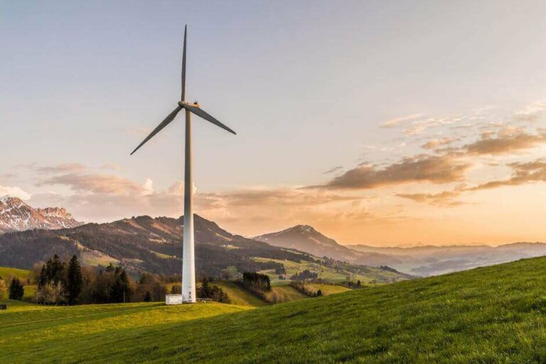 A wind turbine stands in a green, hilly landscape with mountains in the background during a sunset. The sky has shades of orange and yellow, and there are scattered clouds. The scene, an Argentinian renewable energy investment opportunity, is serene and picturesque.