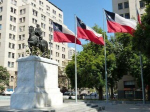 Una estatua ecuestre sobre un pedestal de mármol está situada frente a varias banderas chilenas, simbolizando el espíritu de las elecciones chilenas. La escena se desarrolla en una plaza rodeada de árboles y edificios altos y modernos bajo un cielo azul claro.